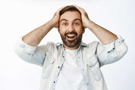 Excited and surprised man cheering, holding hands on head in disbelief and smiling amazed, watching...