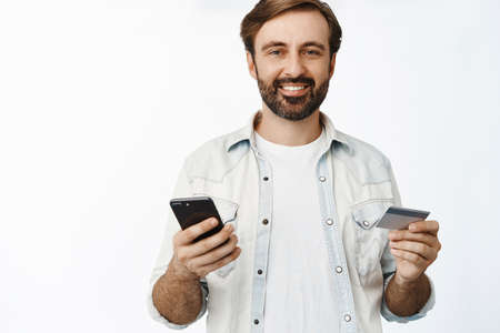 Portrait of handsome guy with credit card and mobile phone, smiling and looking at camera, standing over white backgroundの写真素材