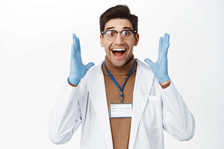 Excited doctor clap hands and smiling amazed, looking ecstatic, standing in medical robe and rubber...