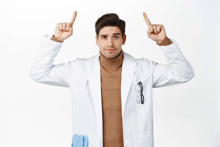 Confused doctor pointing fingers up, looking puzzled, standing in medical robe against white backgroundの写真素材