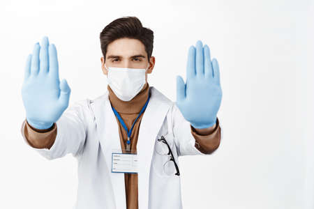 Portrait of mal doctor in face mask and robe, extend hands in gloves, showing stop gesture, taboo disapproval sign, standing over white backgroundの写真素材