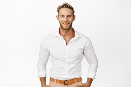 Handsome macho man with blond hair and beard, smiling and looking confident at camera, wearing white shirt, standing against studio backgroundの写真素材