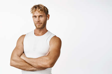 Portrait of handsome sportsman in wireless earphones, cross arms, standing in power pose,...