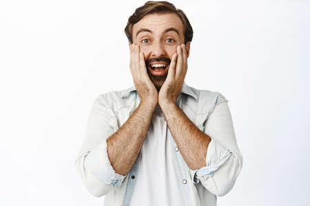 Surprised bearded guy looking in disbelief, smiling amazed, touching his face, standing in casual...