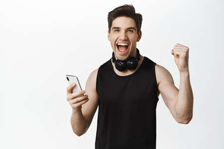 Enthusiastic sportsman with headphones, holding smartphone and shouting of joy, rejoicing, winning, reaching daily goal in mobile app for sport and workout, white backgroundの写真素材