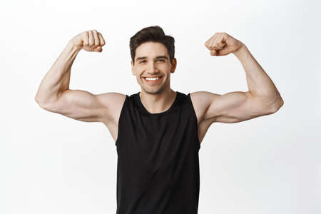 Sport and gym. Smiling young healthy man flexing biceps, showing strong big muscles and fit body, standing in tank top over white backgroundの写真素材