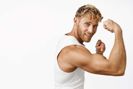 Portrait of handsome fitness athlete flexing biceps, showing strong muscle arm and smiling, workout in gym, standing over white backgroundの写真素材