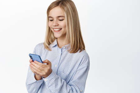 Cute blond girl, teen using mobile phone, chatting or scrolling social media app, standing over white backgroundの写真素材