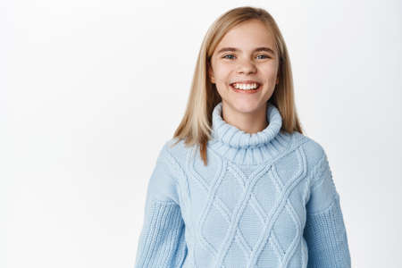 Portrait of happy little girl wearing stylish blue winter sweater, smiling with healthy white teeth, looking at camera, standing over white backgroundの写真素材