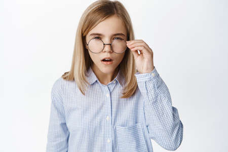 Portrait of blond girl, little kid in glasses squinting, has difficulties reading without eyewear, standing over white backgroundの写真素材