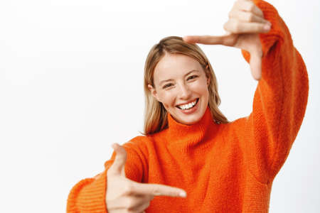 Beautiful woman looking through hand frames, smiling and laughing, imaging, picturing smth, standing in red winter sweater over white backgroundの写真素材