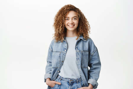 Portrait of modern stylish girl with curly hair smiling, standing in casual outfit. Happy young...