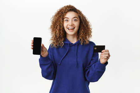 Mobile banking concept. Smiling young woman showing smartphone screen app, credit card, looking...