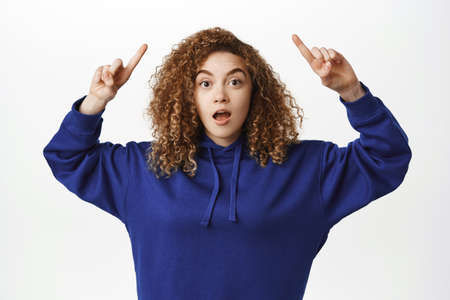 Portrait of smiling curly woman pointing fingers up, showing advertisement on top, looking impressed...