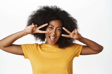 Portrait of happy african american woman shows victory, v-sign peace gesture and smiling, standing in yellow tshirt over white backgroundの写真素材