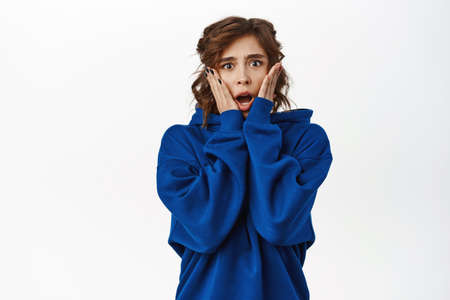 Shocked young woman, horrified, looking scared or worried, standing in blue hoodie over white backgroundの写真素材
