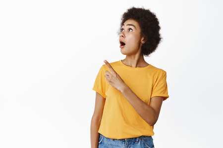 Impressed african girl pointing and looking away at upper left corner direction, showing promo sale, deal in store, standing in yellow tshirt over white backgroundの写真素材
