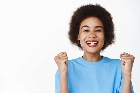 Close up of happy successful african girl, winning and rejoicing, clenching fists and smiling pleased, triumphing, standing in blue tshirt over white backgroundの写真素材