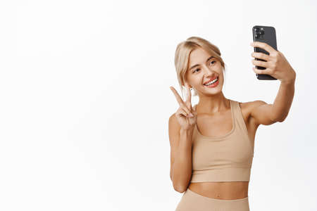 Stylish sport girl in fitness clothing and wireless earphones, taking selfie during workout on smartphone, posing against white studio backgroundの写真素材