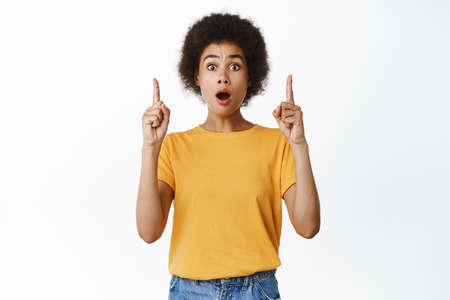 Portrait of beautiful black girl looks surprised, say wow, pointing finger up impressed, showing...