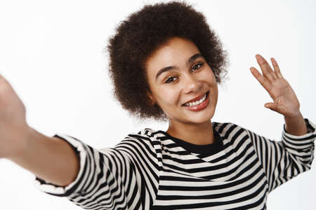 Happy young modern Black girl taking selfie on mobile phone, posing, video chatting on smartphone app, standing over white backgroundの写真素材
