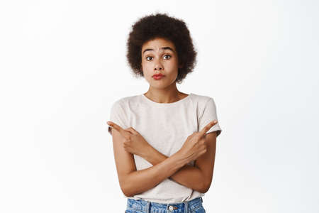 Indecisive cute african girl pointing sideways, showing two variants, choosing between products, standing in tshirt over white backgroundの写真素材