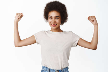 Strength and women workout concept. Smiling black girl with afro hair, young woman flexing biceps,...