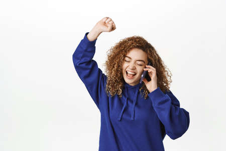 Portrait of smiling young woman talking on mobile phone, having conversation, answer call, standing in blue hoodie over white backgroundの写真素材