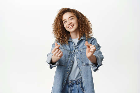Portrait of smiling curly girl, stylish girlfriend shows finger heart, love sign and pucker lips, kissing gesture, standing in denim clothes over white backgroundの写真素材