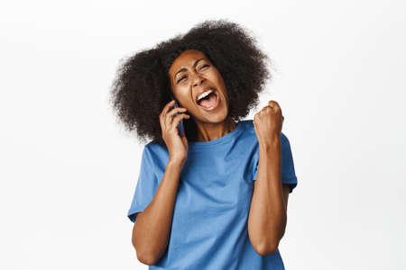 Enthusiastic black woman answer phone call and celebrating victory, hear great news during mobile conversation, standing in blue tshirt over white backgroundの写真素材