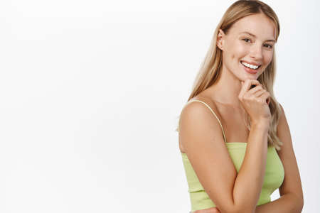 Beauty, skin and hair care. Attractive blond feminine woman with glowing facial skin, no makeup, perfect white smile, posing happy against white background in summer clothesの写真素材