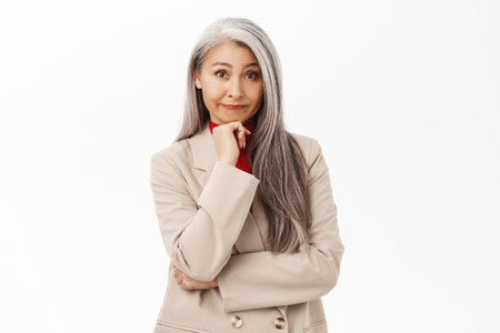 Portrait of korean businesswoman, woman with grey hair thinking, wearing suit, smiling intrigued, standing over white backgroundの写真素材