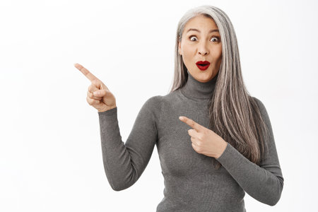 Portrait of excited, surprised asian beautiful lady middle age, pointing fingers left, gasping amazed, showing information, banner or logo, standing over white backgroundの写真素材