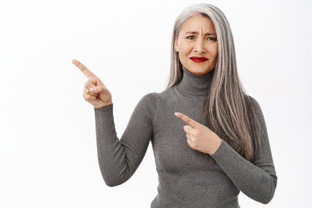 Portrait of disappointed asian senior woman pointing left, grimacing dissatisfied, standing upset against white studio backgroundの写真素材
