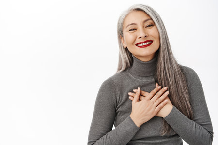 Portrait of asian senior woman, beautiful korean middle aged lady, smiling thankful, holding hand on heart, appreciate, grateful for smth, standing over white backgroundの写真素材