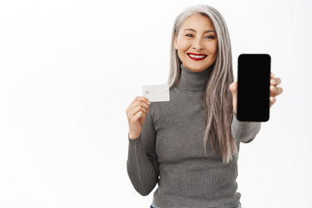 Happy smiling middle aged woman showing mobile phone empty screen and credit card, recommending application, online shopping or bank app, standing over white backgroundの写真素材