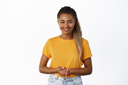 Image of smiling Black woman holding hands together and looking at camera helpful, assisting customer or client, standing friendly against white backgroundの写真素材