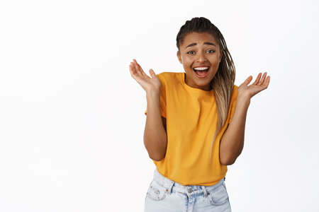 Happy african american girl shrugs and laughs carefree, chuckle over something surprising, standing over white backgroundの写真素材