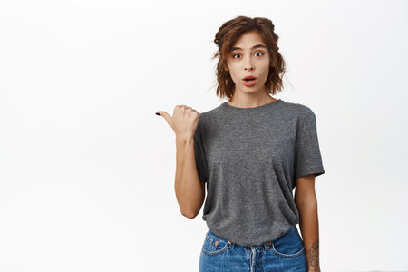 Portrait of girl look surprised when pointing left, showing smth interesting, sharing amazing news, standing in casual clothes over white studio backgroundの写真素材