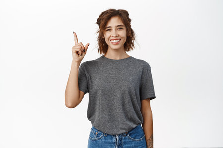 Happy smiling young woman pointing finger up, showing advertisement, top sale promo, standing over white backgroundの写真素材