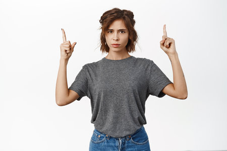 Angry brunette woman complains, points fingers up with frowned upset face expression, stands over white backgroundの写真素材