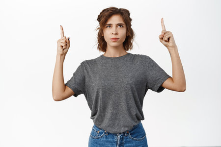 Angry brunette woman complains, points fingers up with frowned upset face expression, stands over white backgroundの写真素材