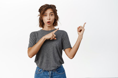 Portrait of surprised brunette woman pointing right, showing advertisement and looking amazed, excited with information, standing over white backgroundの写真素材