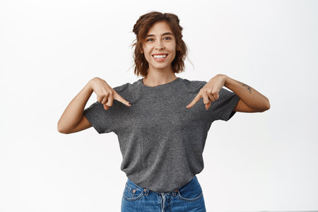 Beautiful smiling girl points fingers down, shows logo, announcement below, standing in casual clothes over white backgroundの写真素材