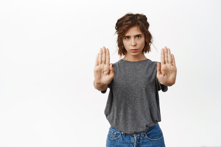 Stop, enough. Serious young woman raising hands block, showing prohibition, warning gesture, dont come closer, stay back, rejecting smth, standing over white backgroundの写真素材