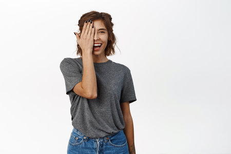 Happy, excited brunette girl with clear facial skin, cover half side of face and looking amazed at camera, standing in grey t-shirt over white backgroundの写真素材
