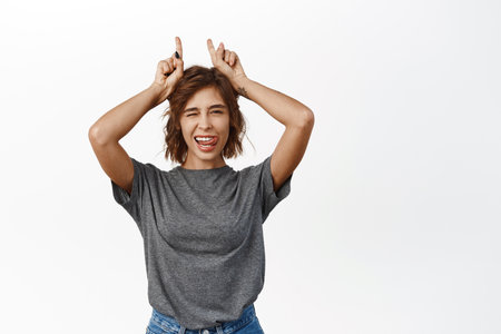 Joyful beautiful girl winking and smiling, showing finger horns, pointing up, standing silly against white backgroundの写真素材
