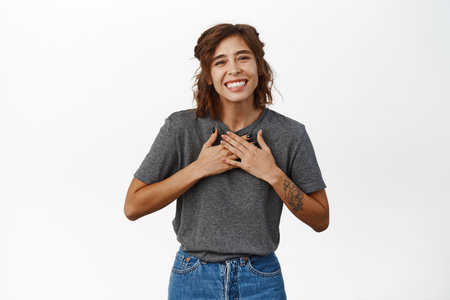 Happy young natural girl, holding hands on chest in appreciation pose, smiling and laughing, receive compliment, praise, say thank you, standing over white backgroundの写真素材