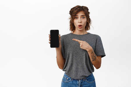 Surprised and amazed young woman, pointing finger at smartphone screen, asking question, showing smth on mobile phone, standing against white backgroundの写真素材