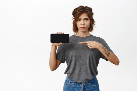 Girl with doubts, skeptical face emotion, pointing at horizontal phone screen, cellphone display, looks concerned, asks question, stands over white backgroundの写真素材